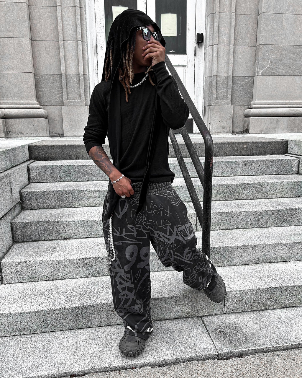 Young adult wearing black vandalizm sweats with graffiti print, posing on steps in urban setting.