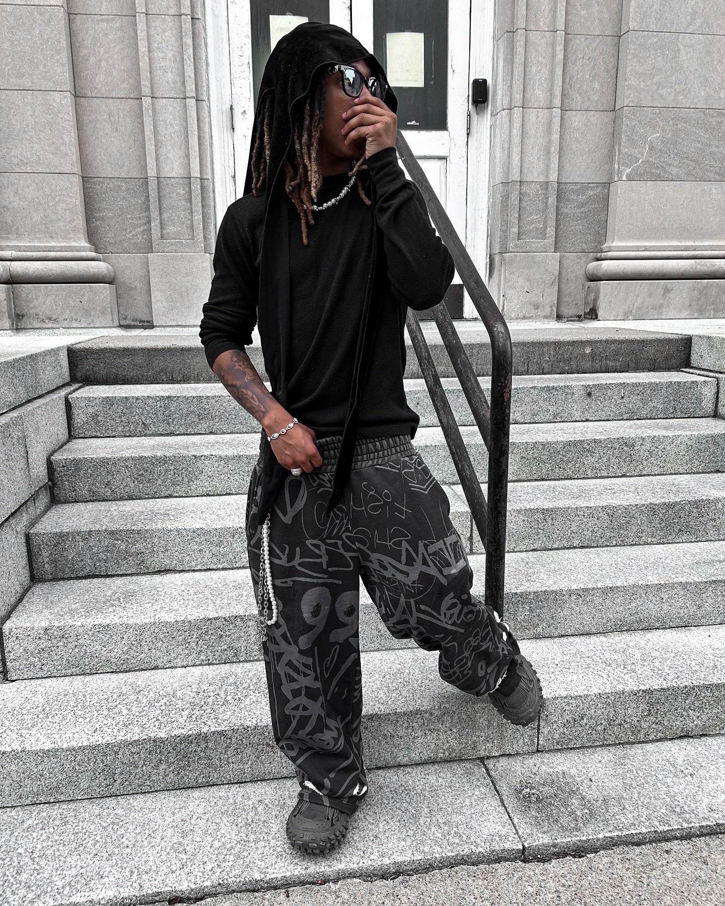 Young adult wearing black vandalizm sweats with graffiti print, posing on steps in urban setting.