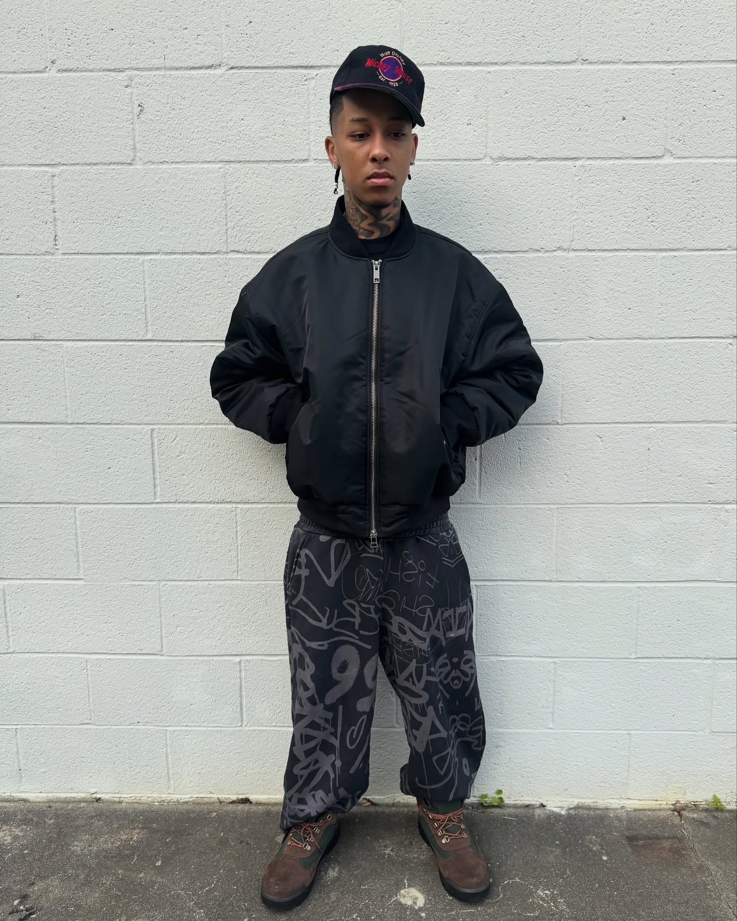Young man wearing a black bomber jacket and patterned black pants, posing against a white wall.